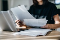A cropped shot of a woman handling paper medical bills while working on a laptop at home.
